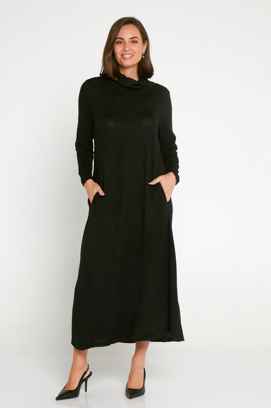 A woman standing in a studio setting, wearing a long black maxi dress with a cowl neck and long sleeves, in a relaxed fit, with the dress extending to the calf. She has her hands in the pockets of the dress.