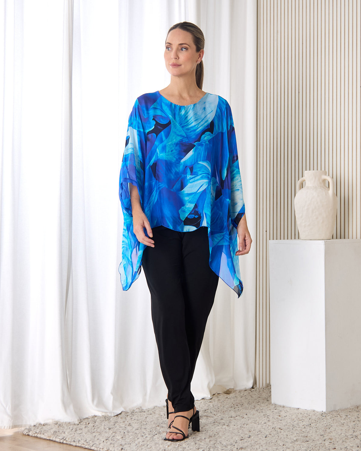 Palermo Silk Top in blue floral, full-length view styled with black pants in a living room setting