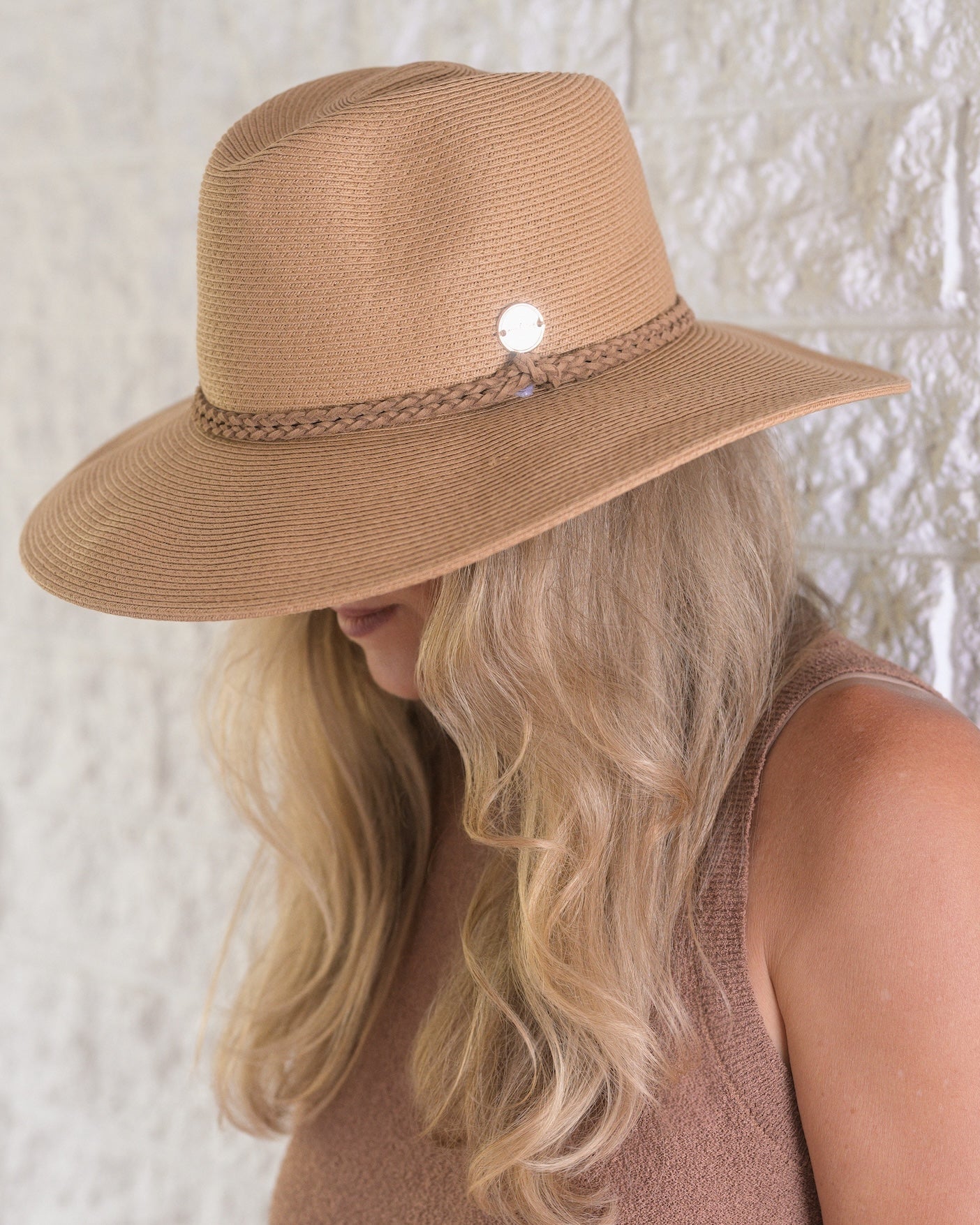 Person wearing a brown straw hat with a decorative band against a textured white wall.