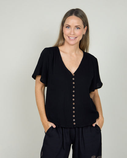 Tarin Button Top in black, front view worn untucked with hands in pockets